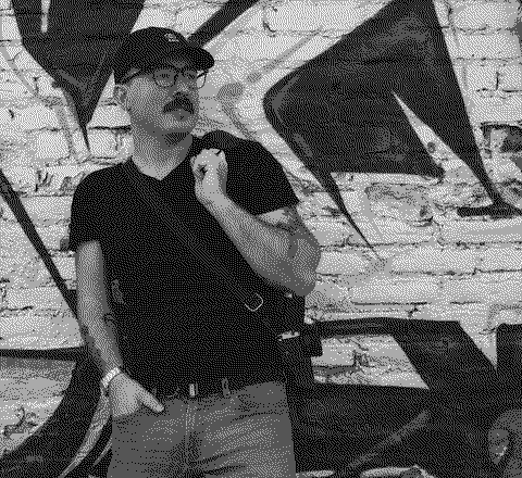 Antonin in a black cap and glasses, leaning against a graffiti-covered brick wall.